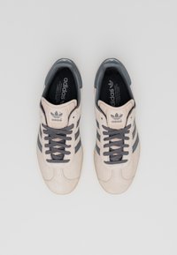 Ljust beige sneakers med mörkgrå ränder, rund tå och snördesign. Ovandel i mocka och gummisula med Adidas-logotyp på tungan och innersulan.
