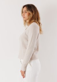 Pullover color beige chiaro realizzato in un materiale morbido e testurizzato, con maniche lunghe e scollatura a V, abbinato a pantaloni bianchi.