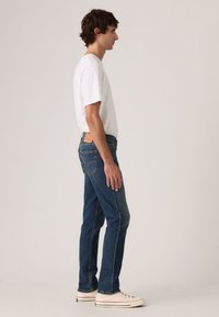 Σκούρα μπλε τζιν denim με στενή εφαρμογή, με δερμάτινο λογότυπο στη μέση και ραφές στο πλάι. Φορεμένα με λευκό t-shirt.