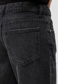 Záber zblízka na zadné vrecko čiernych riflí z denimu s viditeľným prešitím a štítkom "perfect jeans gina tricot" na opasku.