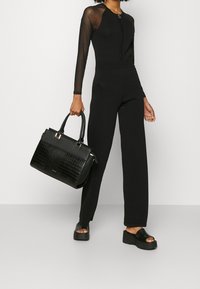 Combinaison noire avec de longues manches transparentes, corsage ajusté et pantalons évasés. Sac à main noir avec un motif crocodile et des accents brillants. Sandales compensées noires.