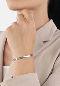 Donna che indossa un sottile braccialetto d'argento lucido al polso, vestita con un blazer beige chiaro e una maglietta bianca sotto.