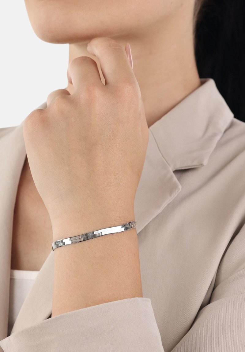 Donna che indossa un sottile braccialetto d'argento lucido al polso, vestita con un blazer beige chiaro e una maglietta bianca sotto.