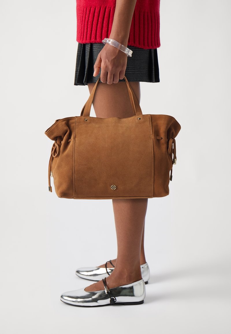 Personne tenant un grand sac à main en daim marron, portant un pull rouge, une jupe noire, un bracelet transparent et des chaussures plates argentées brillantes avec des brides noires.