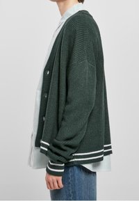 Cardigan verde a maglia con accentuazioni bianco a strisce, caratterizzato da una vestibilità comoda, apertura frontale e polsini a coste. Abbinato a una camicia a righe sotto.