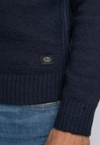Granatowy sweter z dzianiny z prążkowanymi mankietami i dolnym wykończeniem. Posiada czarną metkę z napisem "Pirelli Industries" z przodu na dole. Miękka tekstura.