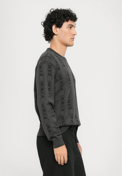 Grauer Strickpullover mit Rundhalsausschnitt, der über ein strukturiertes, vertikales Branding und gerippte Bündchen verfügt, kombiniert mit schwarzen Hosen.