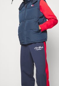 Granatowa puchowa kamizelka z wysokim kołnierzem, bocznymi kieszeniami oraz czerwonymi długimi rękawami pod spodem. Ciemne spodnie dresowe z czerwonym paskiem po boku i białym napisem „New York”.