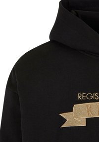 Sweat à capuche noir avec ruban brodé beige et texte partiel "REGIS" visible sur la poitrine, montrant les détails de l'épaule et de la capuche.