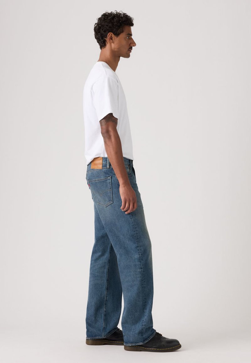 Jeans de mezclilla azul claro con un corte holgado, cintura de tiro medio y una etiqueta marrón en la parte trasera. Combinados con una camiseta blanca lisa y zapatos negros.