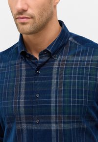 Camicia a quadri blu con colletto a bottoni, realizzata in un tessuto testurizzato e con strisce chiare e scure in contrasto che attraversano tutto il motivo.
