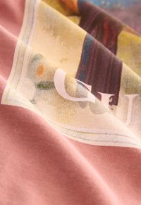 Pink fabric with a partially visible, colorful printed design featuring yellow, green, and dark shapes, shown with soft folds and texture.