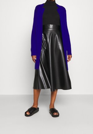 Personne portant un col roulé noir, une jupe midi en cuir noir taille haute, un cardigan ouvert bleu électrique et des sandales slides noires.
