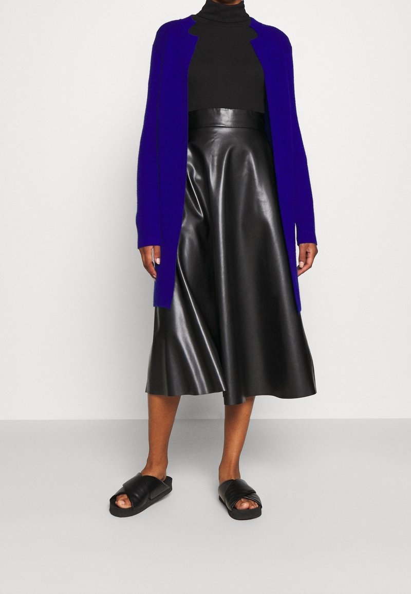 Personne portant un col roulé noir, une jupe midi en cuir noir taille haute, un cardigan ouvert bleu électrique et des sandales slides noires.