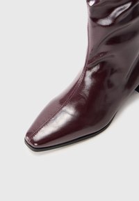 Burgundy leather ankle boot with a pointed toe, shiny finish, and subtle stitching details on the side and front. Black textured sole.