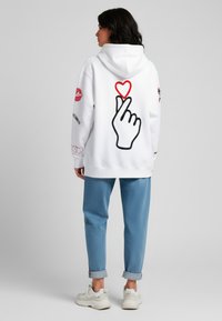 Felpa bianca con una grande grafica di una mano contornata in nero e un cuore rosso sul retro. Abbinata a pantaloni di jeans blu arrotolati e sneakers.