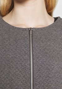 Vêtement gris à fermeture éclair avec une finition texturée. Il présente un col rond et une fermeture éclair en acier inoxydable à l'avant.