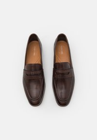 Mocasines de cuero marrón oscuro con un detalle de tira trenzada, que cuentan con un diseño elegante y puntera redondeada. Se puede apreciar una textura suave y costuras sutiles.