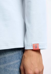 Lichtblauw langemouw shirt met een zachte textuur. Heeft een klein oranje label met een hondenontwerp op de manchette. Eenvoudig gestikte zoom.