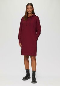 Burgunderfarbendes Hoodie-Kleid aus weichem Stoff, mit langen Ärmeln, einer Kängurutasche und geradem Schnitt, kombiniert mit schwarzen Knöchelstiefeln.