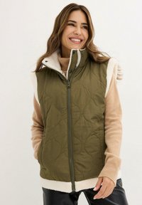 Gilet verde trapuntato con colletto alto, morbida fodera bianca e chiusura con zip. Indossato sopra un maglione beige, presenta tasche frontali e dettagli testurizzati.