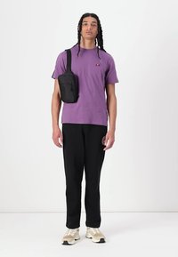 Camiseta de algodón morada con un pequeño logo, pantalones negros con un acento y zapatillas beige. El modelo sostiene un bolso negro de hombro.