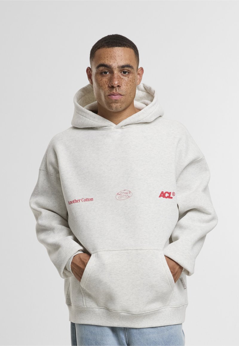 Ung mann med kort hår og fregner har på seg en lys grå hettegenser med rød tekst "Another Cotton" og lyseblå jeans, hender i frontlommen.