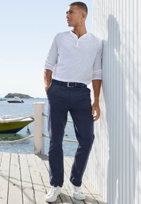 Marineblaue Chinos mit geradem Schnitt, kombiniert mit einem hellgrauen Langarm-Henley-Shirt und weißen Sneakers, stehen an einer Holzwand.