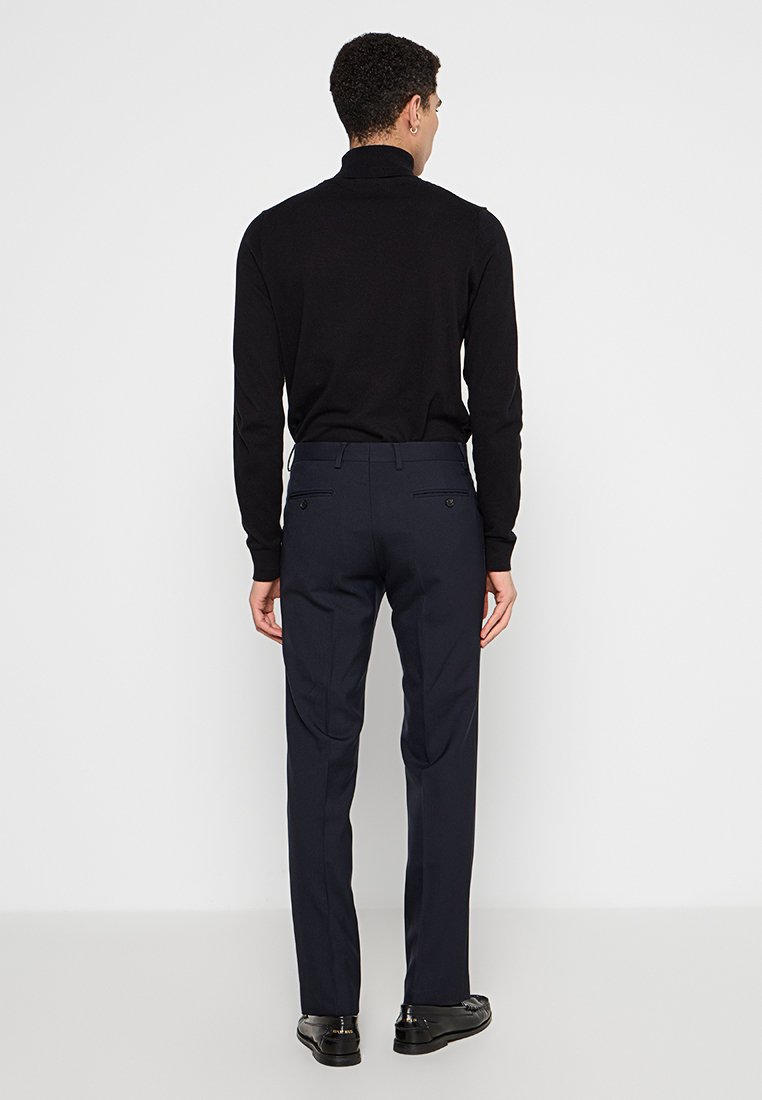 Suéter negro de corte ajustado con cuello alto; pantalones de vestir azul marino con bolsillos frontales y traseros; modelo de espaldas, de pie sobre un fondo liso.