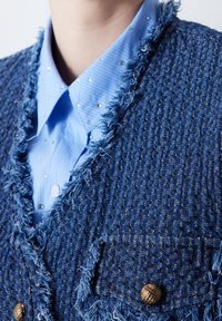 Blazer en tweed bleu avec des franges, doté de boutons en métal doré et d'un motif texturé. Associé à une chemise bleu clair ornée de strass.