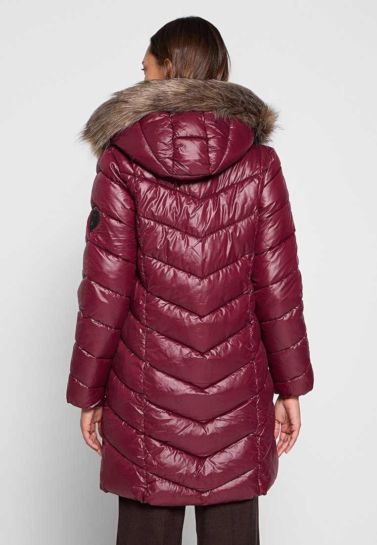 Vrouw in een lange, gewatteerde burgundy winterjas met een met bont afgewerkte capuchon, staand met de rug naar de camera tegen een effen achtergrond.