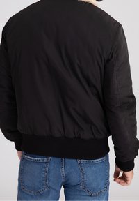 Svart bomberjacka med fuskpälskrage, har ribbad nederkant och mudd, samt slät tygtextur. Bärs med blå jeans, visar bakvy.