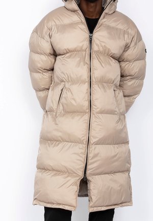 Homme portant un long manteau matelassé beige avec fermeture éclair à l'avant et poches latérales zippées, mains dans les poches.