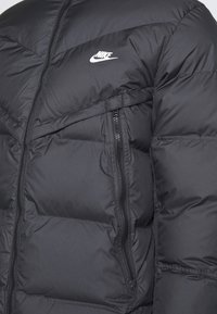 Svart vadderad Nike-jacka med framdragkedja, dragkedjeförsedd sidoficka och vitt Nike-logotyp på bröstet.