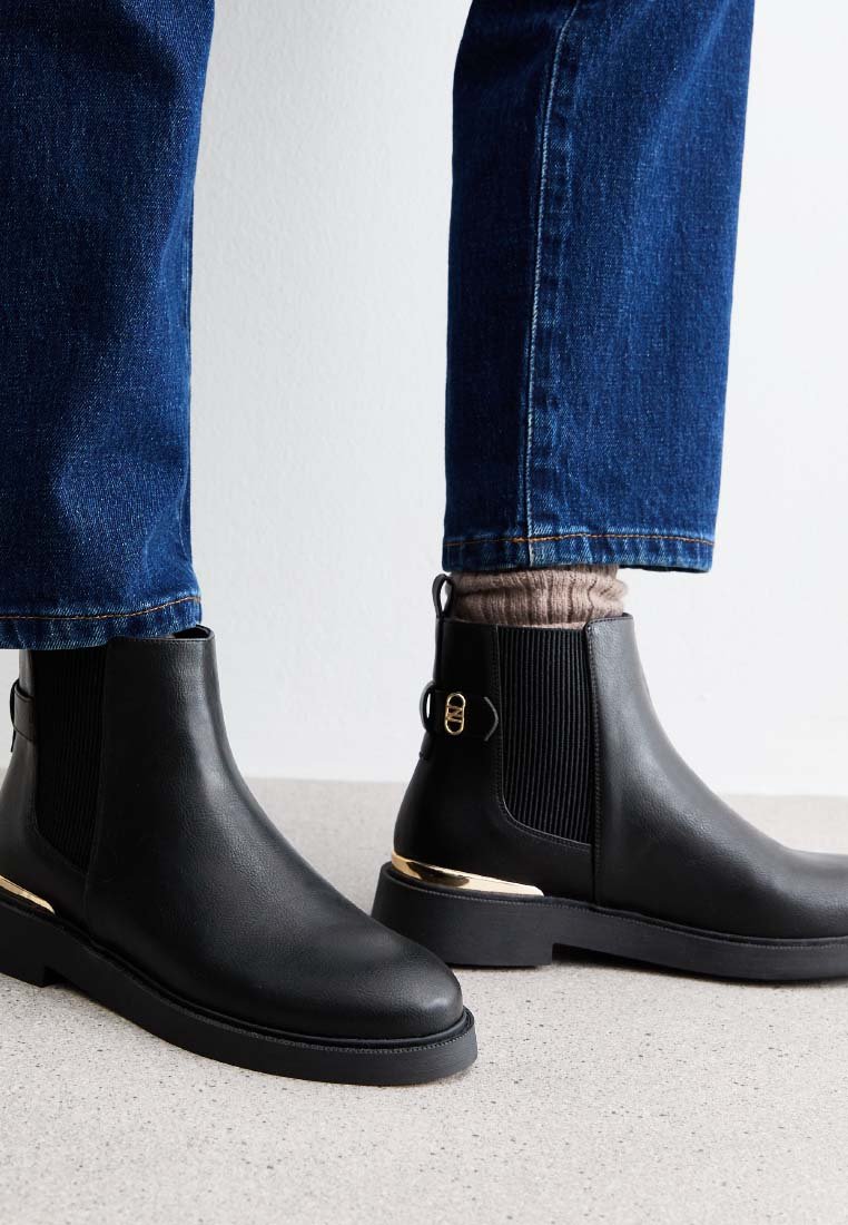 Black leather ankle boots with elastic side panels and a gold accent on the heel, paired with blue denim jeans and a textured gray sock.