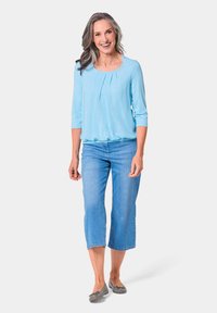 Hellblaue Bluse mit 3/4-Ärmeln und Faltendetail, kombiniert mit 7/8-Denim-Jeans. Graue Ballettschuhe runden das Outfit ab.