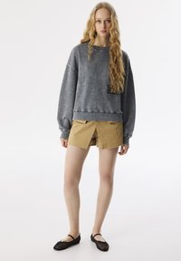 Felpa grigia con finitura texturizzata e una tasca a toppa nera, abbinata a pantaloni corti khaki con un design plissettato e orli grezzi. Ballerine nere.