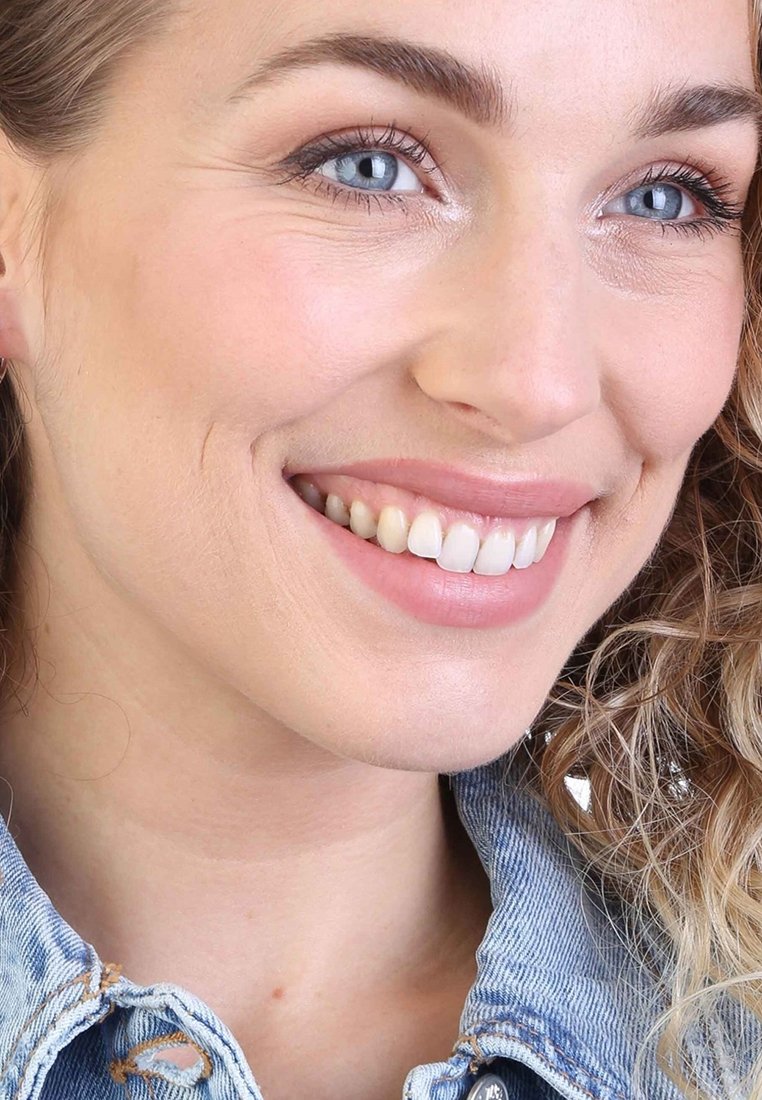 Close-up van een glimlachend gezicht met blauwe ogen, zichtbare lichte huidtextuur, roze lippen en krullend haar, gekleed in een spijkerjack.