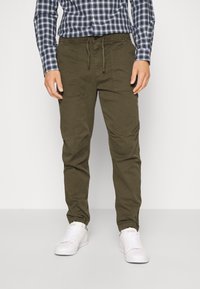 Pantalones cargo verde oliva con cintura ajustable, bolsillos laterales y un diseño de pierna ajustada, combinados con una camisa de cuadros y zapatillas blancas.