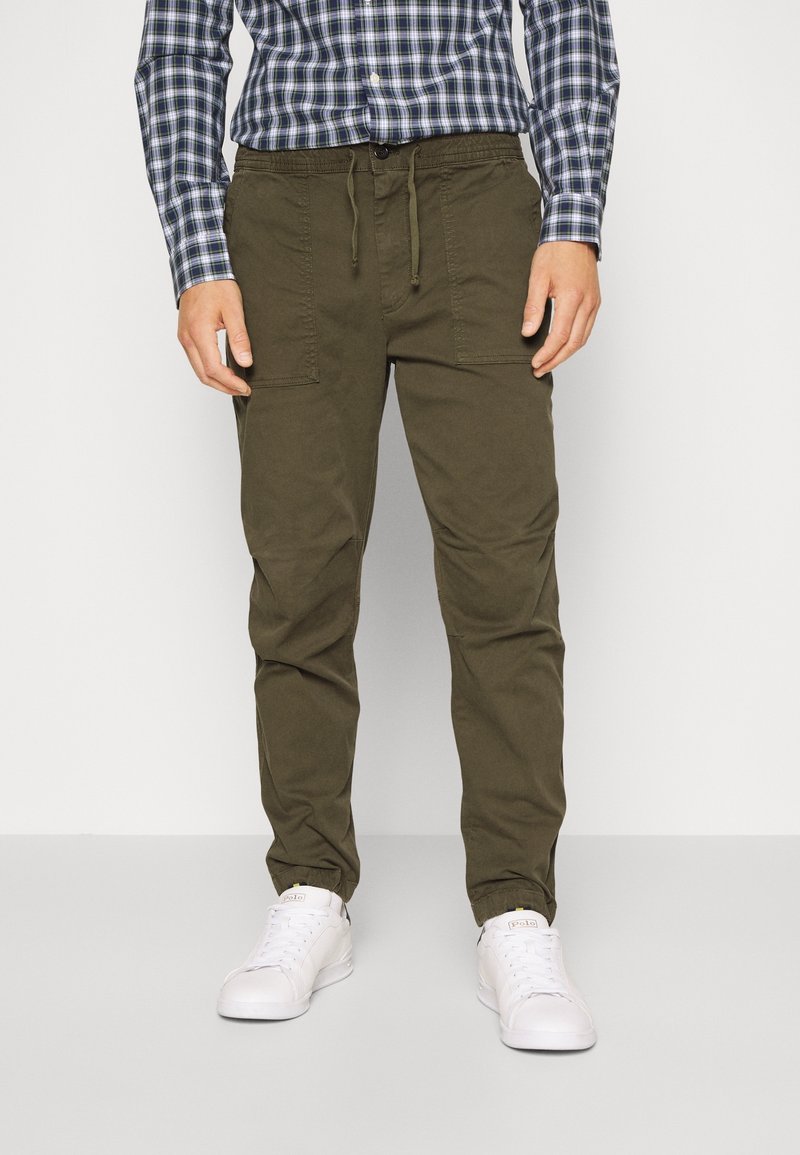 Pantalones cargo verde oliva con cintura ajustable, bolsillos laterales y un diseño de pierna ajustada, combinados con una camisa de cuadros y zapatillas blancas.