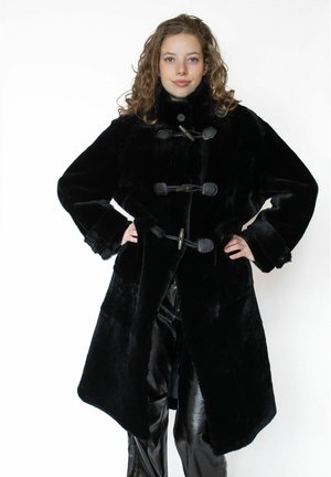 Nuori nainen, joka pukeutunut mustaan tekoturkistakkiin, jossa on kiinnityskoukut, ja mustiin kiiltäviin housuihin, seisoo käsivarret lanteilla valkoista taustaa vasten.