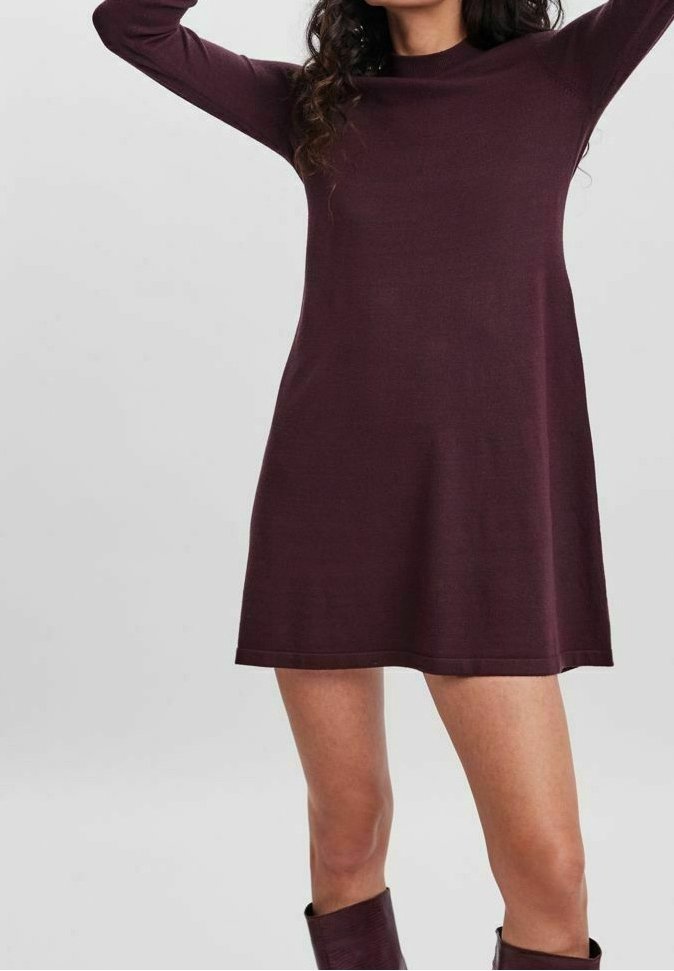 Femme portant une robe prune à manches longues et longueur genou, associée à des bottes prune assorties, debout les bras levés devant un fond gris uni.