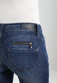 Mörkblå denim jeans med en ficka med dragkedja, broderade sömmar och en märkesläderetikett med nitar på midjebandet.