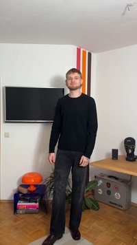 Camisola preta de mangas compridas, jeans escuros com desenhos bordados, sapatos engraxados, numa sala minimalista com uma televisão e detalhes coloridos.