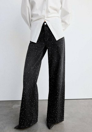Pantalones negros de pierna ancha con patrón moteado blanco, llevados con una camisa blanca abotonada y zapatos negros puntiagudos sobre un suelo gris.