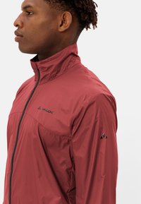 Giovane in profilo che indossa una giacca Vaude bordeaux con zip, colletto alto e logo sul petto e sulla manica, su sfondo bianco.