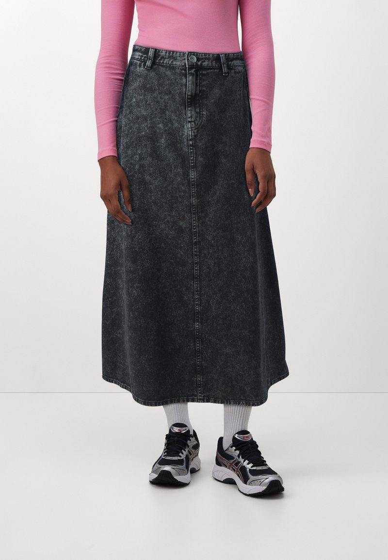 Personne portant une jupe midi en denim noir effet acidulé, un haut rose à manches longues, des chaussettes blanches et des chaussures de sport noires et blanches, debout.