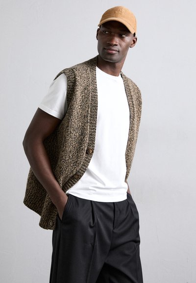 Gilet lavorato in tonalità marrone sopra una t-shirt bianca, con scollatura rotonda e bottoni. Il modello indossa pantaloni neri e un berretto beige.