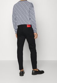 Uomo che indossa pantaloni neri slim-fit con etichetta rossa "HUGO", mocassini neri e una camicia a maniche lunghe con un motivo astratto in bianco e nero.
