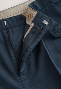Pantalon bleu marine avec une fermeture éclair visible, un bouton et une ceinture plate. Le tissu a une texture lisse, avec des accents beige à la taille.